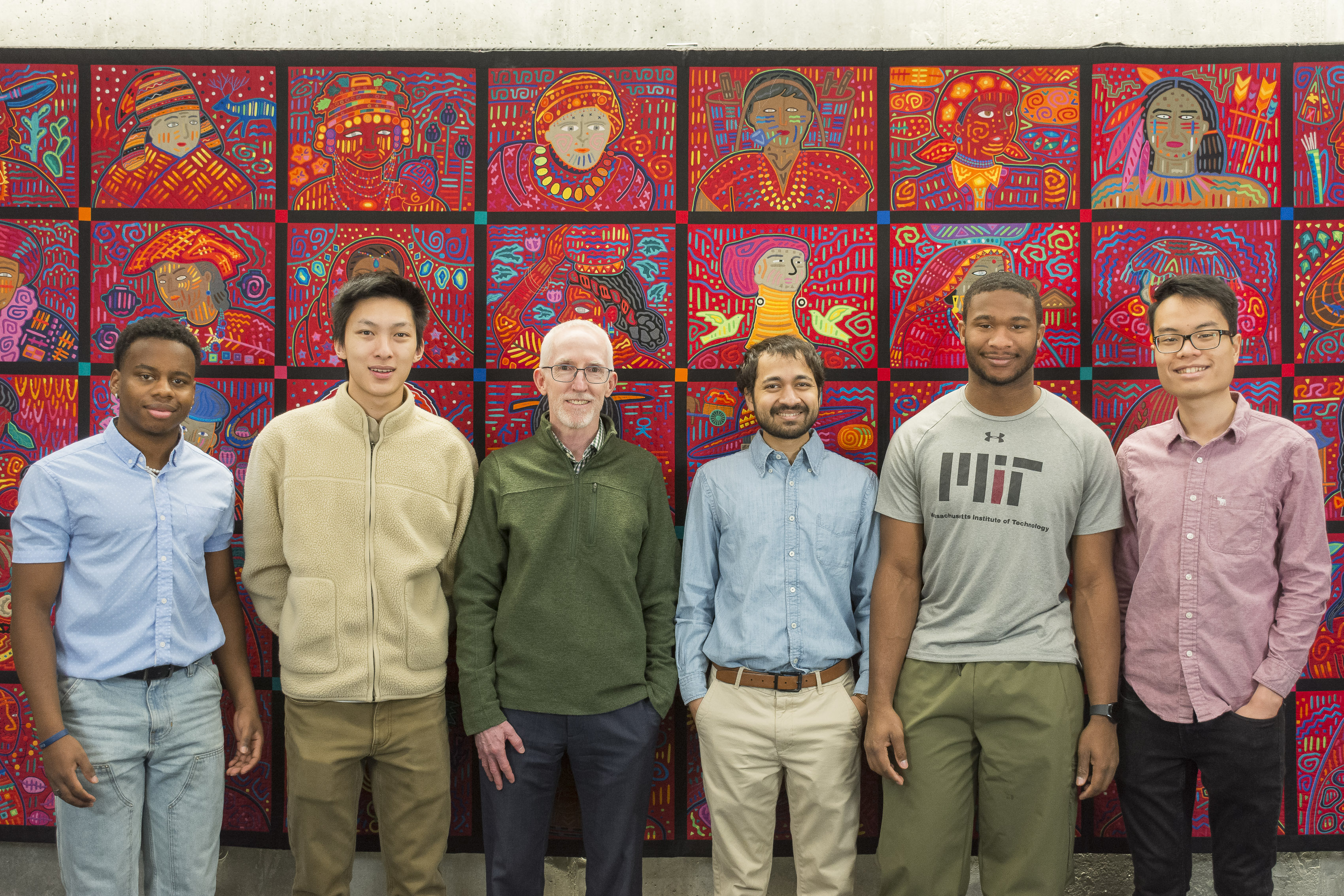 Six Members of the Doyle Group stand against a red tapestry with different faces. The people are: undergraduate students Andre Hamelberg and Ian Chen, Professor Patrick Doyle, Devashish Gokhale PhD '24, and undergraduate students Andrew Okyere and Dien Nguyen. 