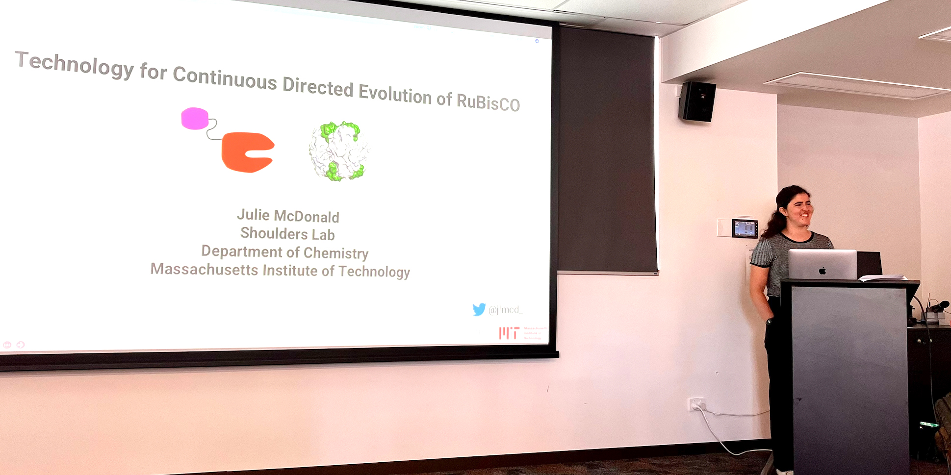 Julie McDonald stands at a podium giving a presentation on Technology for Continuous Directed Evolution of RuBisCO