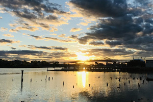 Photo of harbor at sunset with clouds in the sky. There is a bridge on the water in the distance.