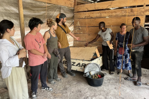 Seven people standing by a wooden box and a load of beeswax thermal batteries in a rough structure with a dirt floor