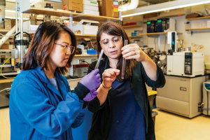 Desirée Plata, right, is wearing a blue lab coat and goggles while showing her grad student, left, a liquid in a test tube. This is taking place in a laboratory. 