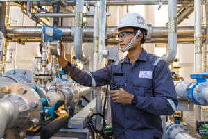 An image of a man in a blue shirt, white hard hat, and work goggles examining a water purification system consisting of piping. 