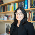 Tavneet Suri in a black blazer and glasses smiling in front of a bookshelf full of books